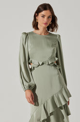 A woman is wearing a light green dress with long, puffy sleeves and a ruffled detail around the waist and hem. The dress has a smooth, satin-like fabric with a slight sheen, and the ruffles add a decorative touch to the overall elegant look. She has shoulder-length brown hair and is accessorizing with gold earrings.